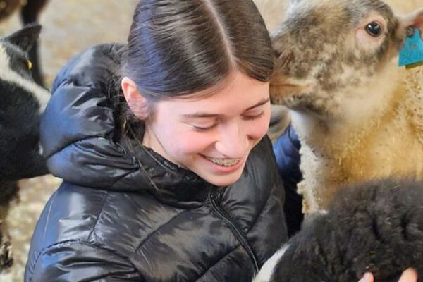 Leende ung kvinna i svart dunjacka klappar ett lamm med gul öronbricka medan ett annat lamm nosar vid hennes kind. Scenen utspelar sig i en fårhage eller ladugård med halm i bakgrunden.