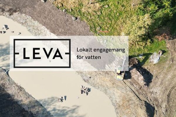 Flygvy över en utgrävd damm med ljus sandig botten där en grävmaskin arbetar vid kanten, omgiven av gräs och träd. Överlagrad text: “FLEVA” och “Lokalt engagemang för vatten”.