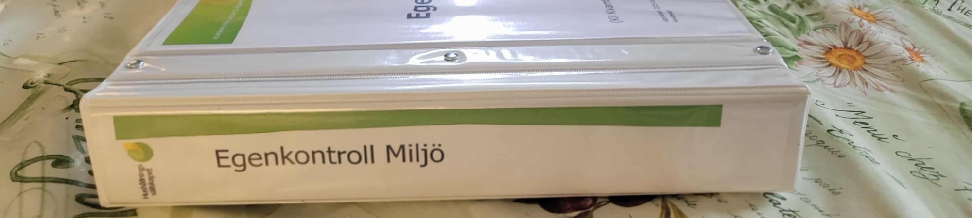 Vit pärm ligger på ett blommigt bord och visar ryggtexten ”Egenkontroll Miljö” med grön markering; på sidan syns en liten logotyp och texten ”Hållbarhet Gotland”.