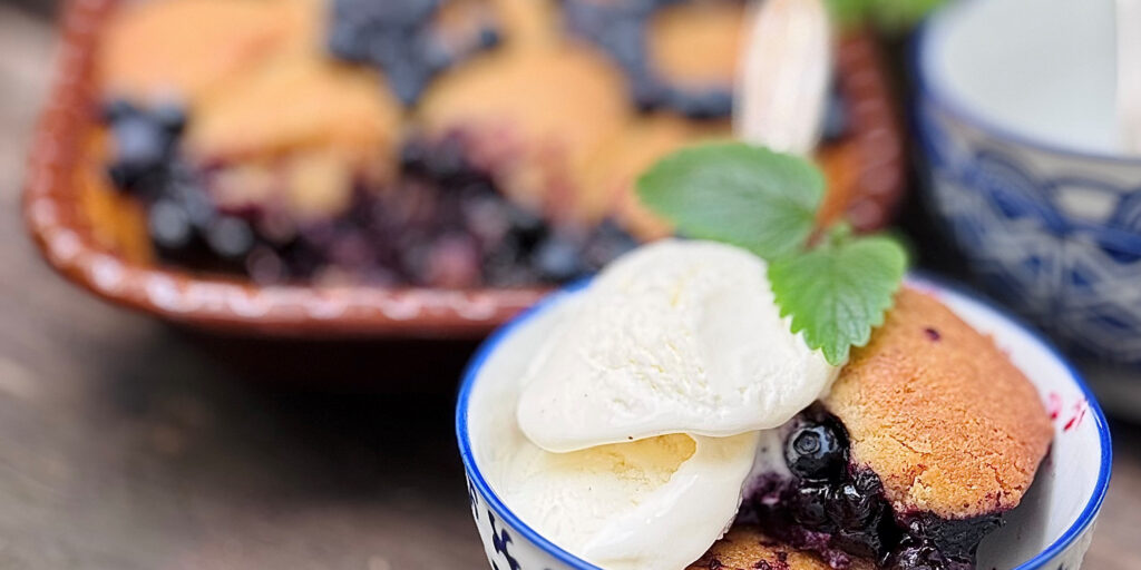 Blåmönstrad skål med blåbärspaj serveras med två kulor vaniljglass och en grön myntakvist ovanpå. I bakgrunden syns en ugnsform med mer paj och en blåvit kopp, lätt ur fokus.