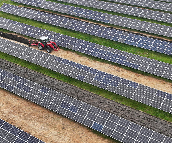 Drönarfoto över demoanläggning för solbruk i Varberg. Röd traktor kör längs en serviceväg mellan långa rader av solpaneler och verkar sköta underhåll i en solcellspark, sedd uppifrån med parallella panelrader och gräsremsor mellan.