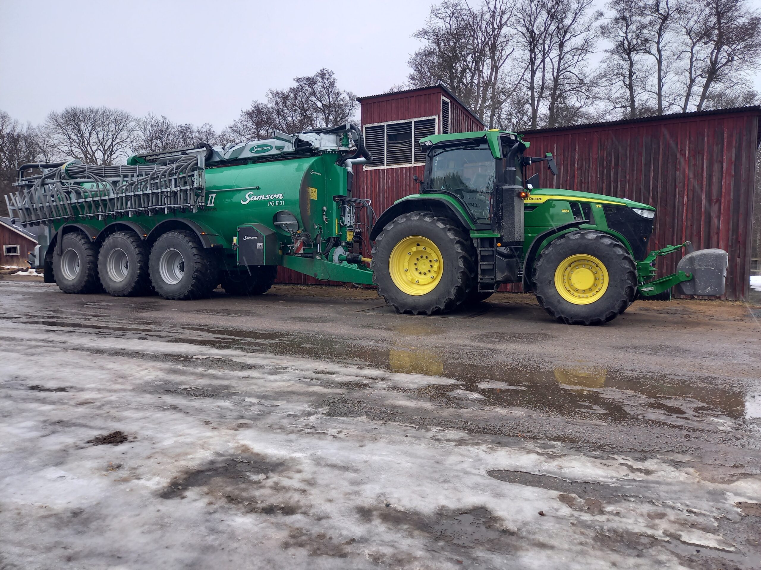 Grön John Deere-traktor står parkerad med stor gödseltankvagn bakom. Tankvagnen har slangar och texten “Samson PG II 31”. I bakgrunden syns en röd ladugård och kala träd, med is och slask på marken.
