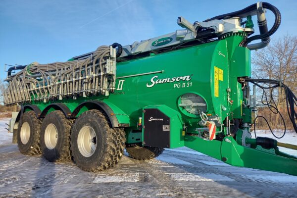 Stor grön lantbruksmaskin märkt “Samson PG II 31” står parkerad på en isig gårdsplan. Maskinen har tre stora hjulaxlar och ett fällbart aggregat med många slangar och munstycken, under klarblå himmel.