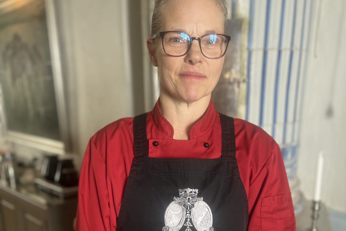 Kvinna i röd kockjacka och svart förkläde står med händerna knäppta och ser in i kameran. Förklädet har en ugglelogga och texten “Bjärsjölagårds Slott”. Bakom syns ett ljust kök.
