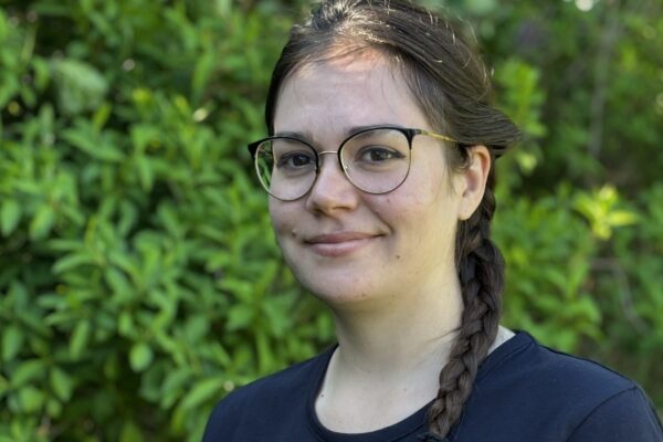 Young woman with glasses and a long braid smiles while standing outdoors in front of leafy greenery. She wears a navy T‑shirt printed “Fältförsök Josefine” and a green leaf logo on the chest.