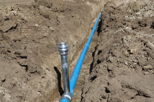 Blå vattenledning ligger nedlagd i en lång, nygrävd schakt genom åkermark, med ett vertikalt rör som sticker upp i förgrunden. Jordvallar kantar diket och gårdar syns långt bort under klar himmel.