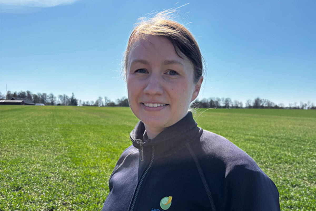 Leende person i mörk jacka står och tittar mot kameran. I bakgrunden syns en stor grön åker med trädlinje vid horisonten under klarblå himmel, i starkt dagsljus.