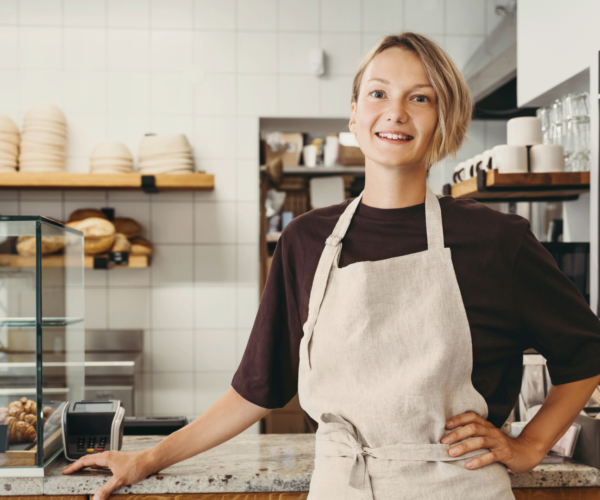 Leende caféanställd i ljus förkläde står bakom disken med handen på höften och lutar sig mot bänken. I bakgrunden syns hyllor med bröd och tallrikar samt en glasmonter med croissanter.