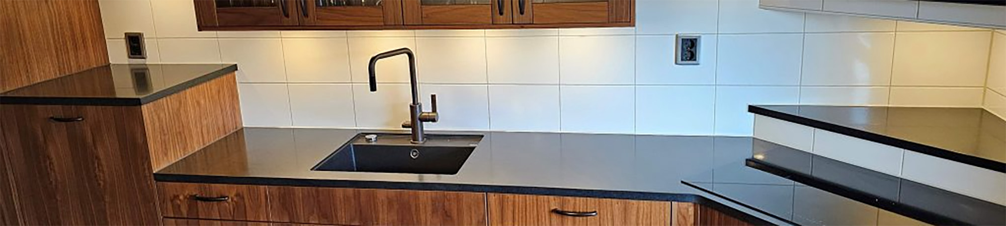 Köksbänk med mörk bänkskiva och infälld svart diskho med blandare, omgiven av träluckor. Bänken löper i vinkel mot en mindre avsats, med vit kaklad vägg och eluttag i bakgrunden.