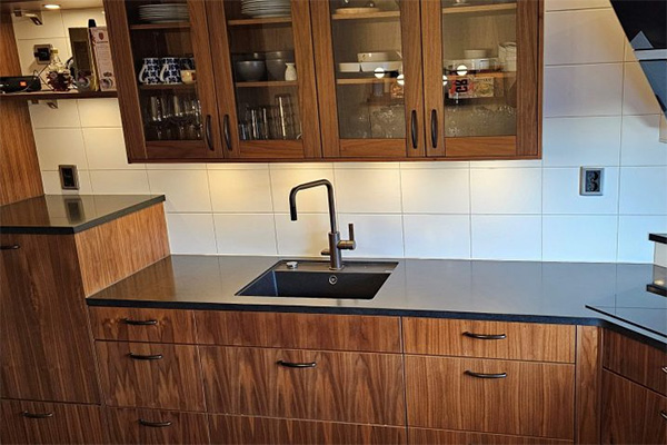 Köksinredning i mörkt trä med bred svart bänkskiva och infälld diskho. Överskåp med glasluckor visar glas och porslin, medan modern svart blandare står centralt mot vit kakelvägg och varma spotlights.