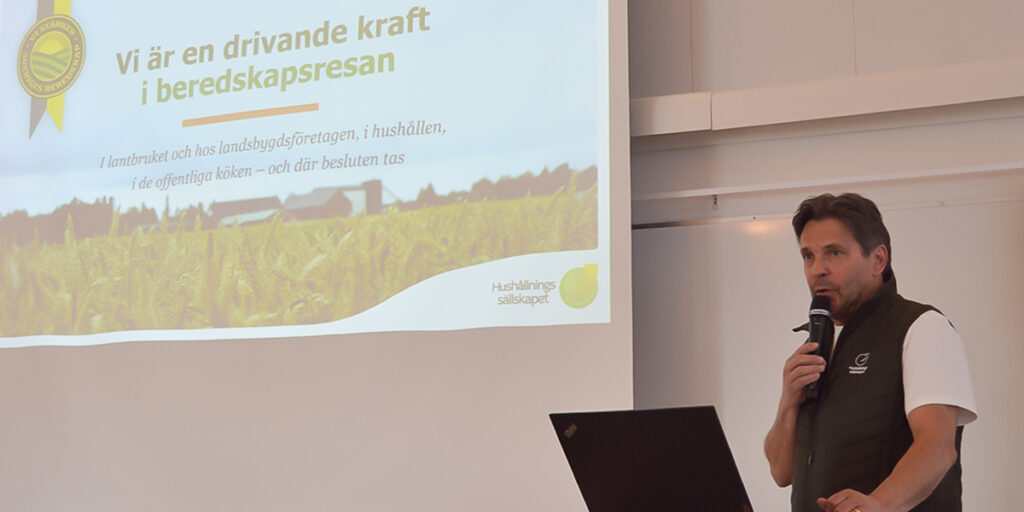 Man står och talar i mikrofon vid en presentation, bredvid en projektorduk. Bilden visar en slide med texten: “Vi är en drivande kraft i beredskapsresan… Hushållningssällskapet” och en bakgrund av åker och landsbygd.