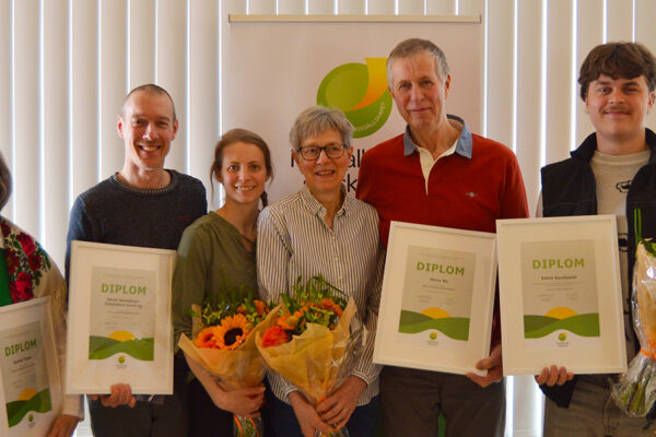 Sex personer står uppradade och ler vid en prisutdelning, håller inramade diplom och blombuketter. Diplomtexten visar rubriken ”DIPLOM”. I bakgrunden syns en rollup med logotyp samt persienner framför fönster.