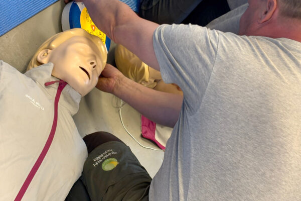 Person placerar hjärtstartarens elektroder på en HLR-docka som ligger på golvet. Close-up av händer vid dockans huvud och en AED-enhet; utbildningsmiljö för säkerhets- och livräddningsträning.