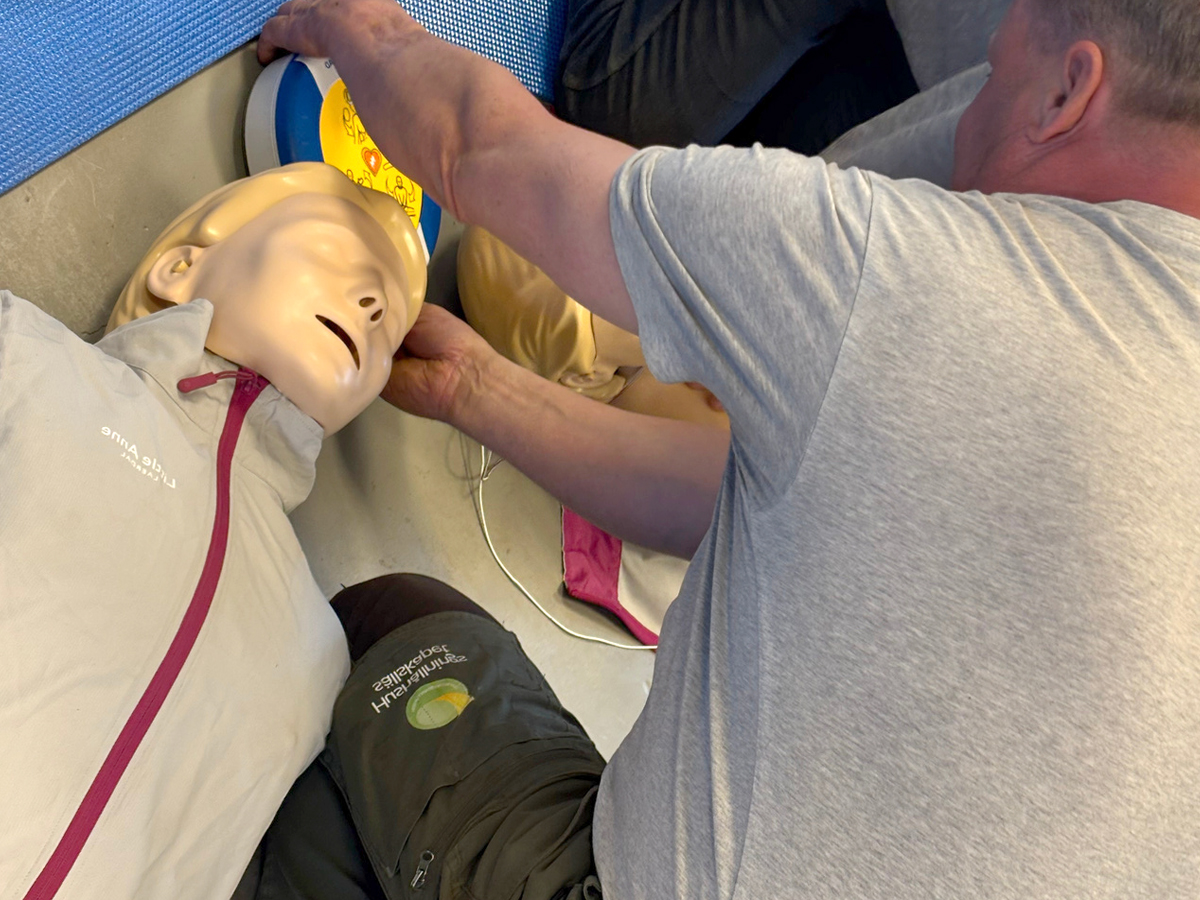 Person placerar hjärtstartarens elektroder på en HLR-docka som ligger på golvet. Close-up av händer vid dockans huvud och en AED-enhet; utbildningsmiljö för säkerhets- och livräddningsträning.