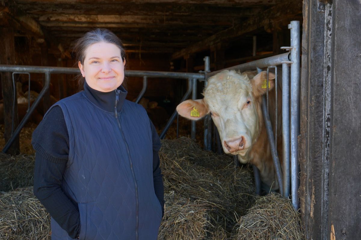 Kvinna i mörk väst står och ler i ett stall, med händerna i fickorna. Bredvid tittar en ljusbrun ko med öronmärken fram genom metallgrindar, omgiven av halm och stallinredning.