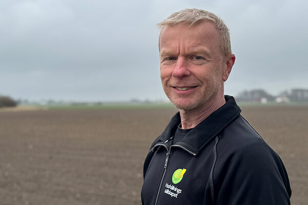 Man med kort hår och mörk jacka står utomhus och ler mot kameran, med en nysådd åker och låga byggnader i bakgrunden under grå molnhimmel. Jackan har texten “Hushållningssällskapet”.