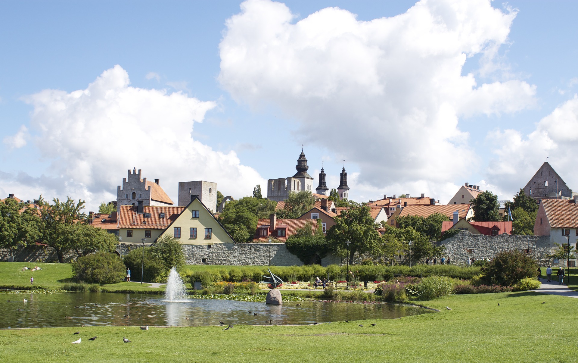 Stadsvy över Visby med damm i förgrunden där en fontän sprutar vatten och fåglar simmar. Besökare promenerar i parken, medan ringmuren, röda tak, kyrktorn och ruiner reser sig under stora vita moln.