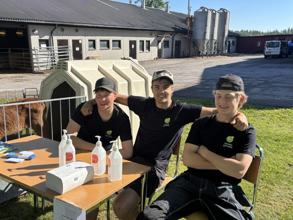 Tre unga män i svarta tröjor med texten “Gamleby gymnasium” sitter vid ett bord utanför en ladugård; en har armen runt de andra och de ler. På bordet står handspritflaskor och pappershanddukar.