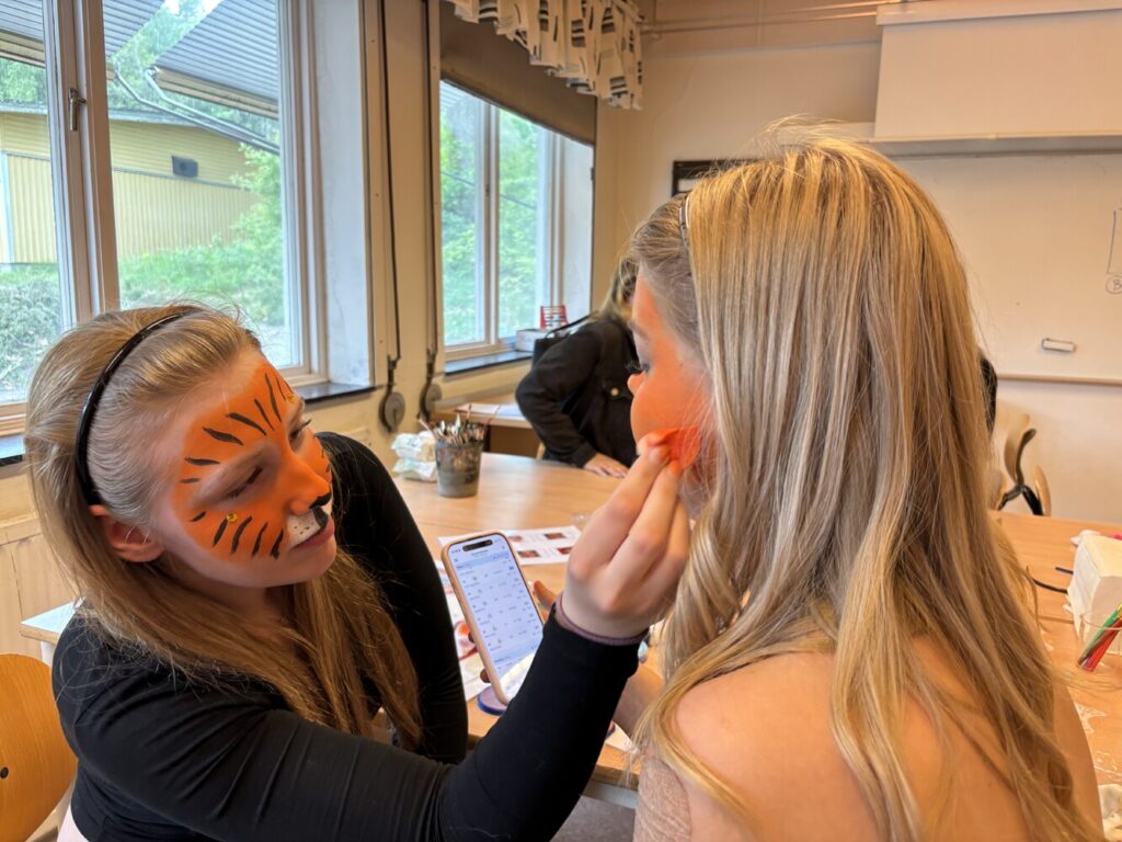 En kvinna med orange tigeransiktsmålning sminkar en annan kvinna genom att dutta rouge på kinden, medan hon håller en mobil med kalender öppen. De sitter vid ett bord i ett klassrum med stora fönster.