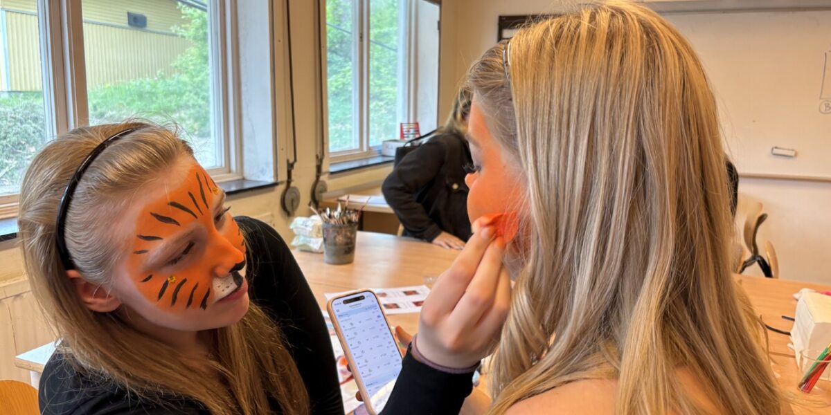 En kvinna med orange tigeransiktsmålning sminkar en annan kvinna genom att dutta rouge på kinden, medan hon håller en mobil med kalender öppen. De sitter vid ett bord i ett klassrum med stora fönster.