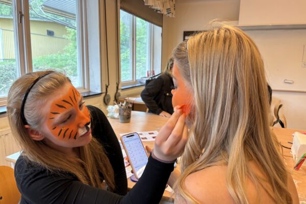 En kvinna med orange tigeransiktsmålning sminkar en annan kvinna genom att dutta rouge på kinden, medan hon håller en mobil med kalender öppen. De sitter vid ett bord i ett klassrum med stora fönster.