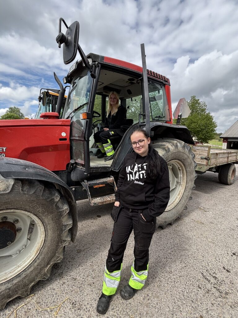 Två personer poserar vid en röd traktor med släp; en står framför med glasögon och hoodie med texten “INATI INATI”, medan den andra sitter i hytten. Utomhus på gårdsplan under molnig himmel.