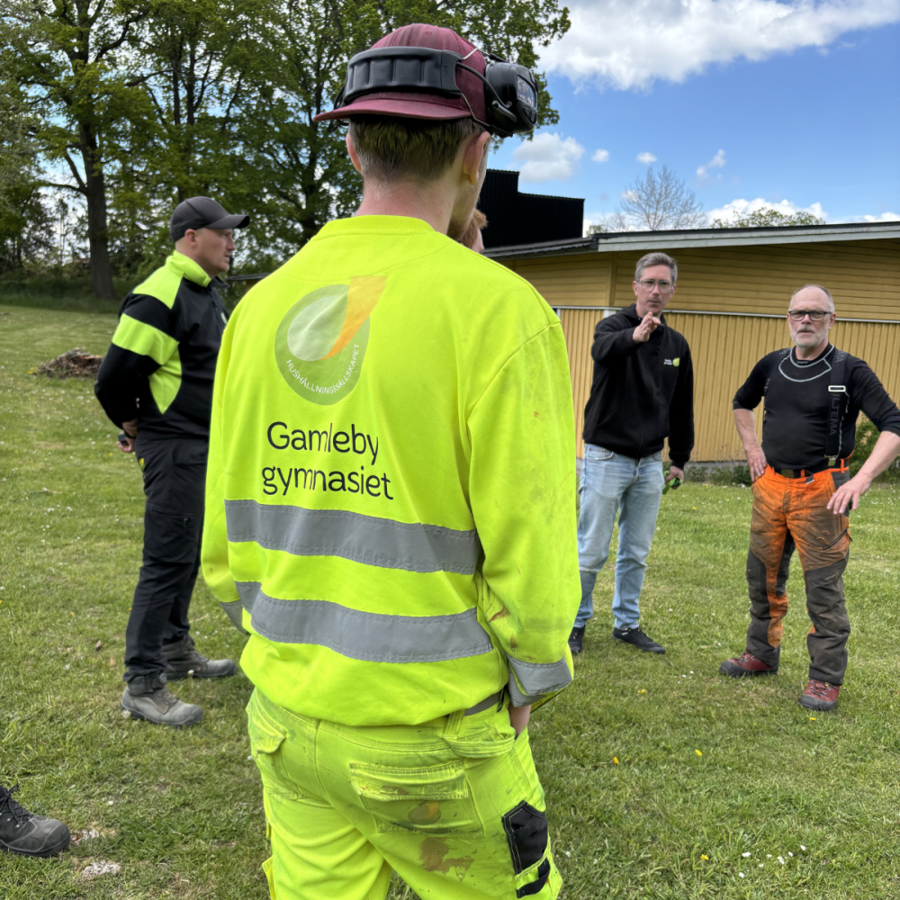 Elev i fluorescerande arbetskläder med texten “Gamleby gymnasiet” står med ryggen mot kameran och lyssnar när en man pekar och instruerar en grupp utomhus på en gräsmatta vid en byggnad.