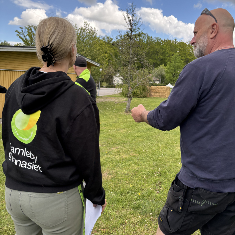 Kvinna i svart hoodie med texten ”Hamleby gymnasiet” och en man står på en gräsmatta och samtalar; mannen gestikulerar medan de tittar mot en tredje person. I bakgrunden syns träd, byggnad och halmbalar.