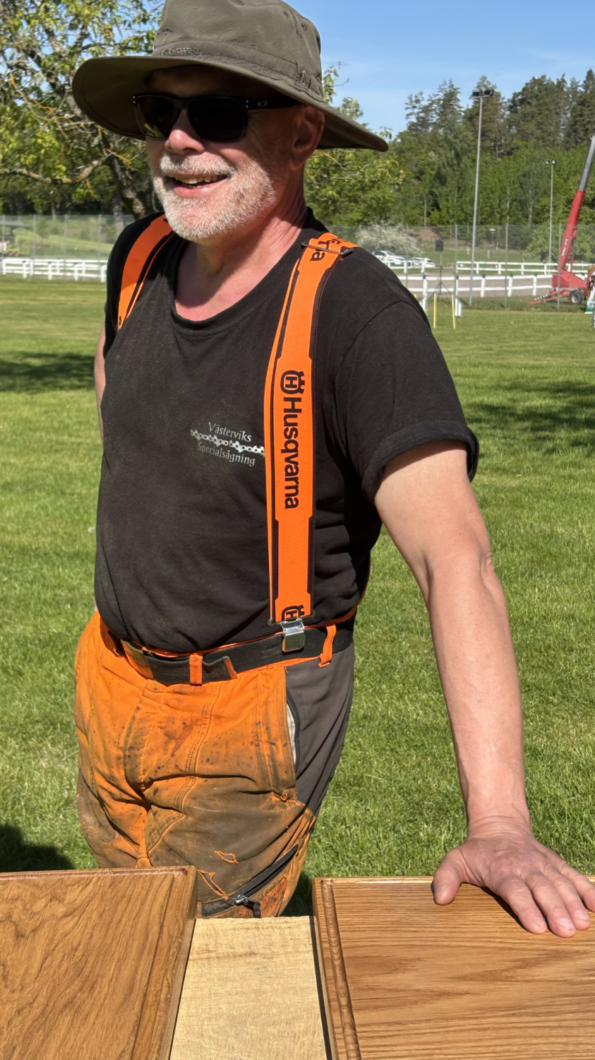 Leende man i solhatt och solglasögon lutar sig mot ett bord med träprover utomhus på en gräsmatta. Han bär orange hängslen med texten ”Husqvarna” och en svart tröja med ”Västerviks Specialsågning”.