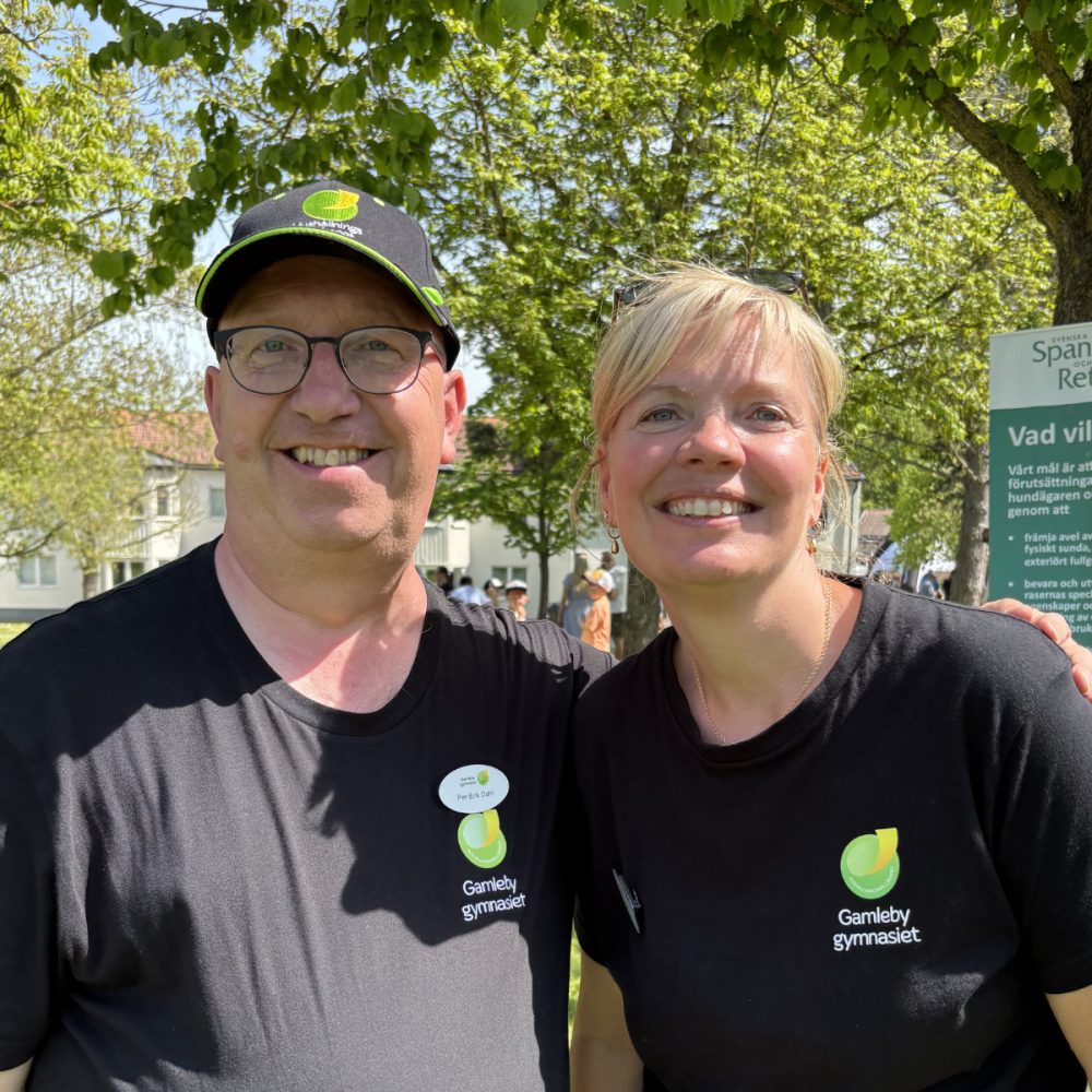 Två personer ler och poserar tätt tillsammans utomhus i en grönskande park. De bär svarta tröjor med texten ”Gamleby gymnasiet” och en grön logotyp; mannen har keps och namnbricka.