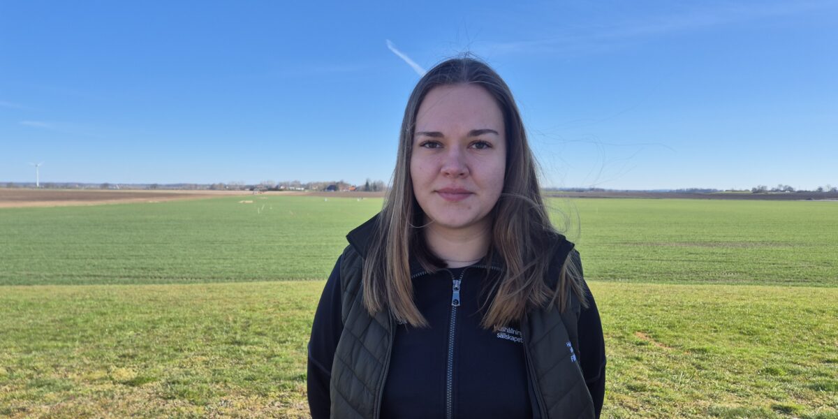 Kvinna med långt brunt hår står och tittar in i kameran på ett öppet jordbruksfält. Hon bär mörk väst och jacka med texten ”Hushållningssällskapet”. I bakgrunden syns gröna åkrar, gårdar och en vindkraftverk under klarblå himmel.