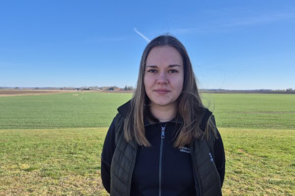 Kvinna med långt brunt hår står och tittar in i kameran på ett öppet jordbruksfält. Hon bär mörk väst och jacka med texten ”Hushållningssällskapet”. I bakgrunden syns gröna åkrar, gårdar och en vindkraftverk under klarblå himmel.
