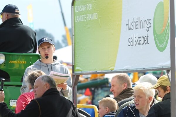 Man med mikrofon och papper står vid en grön John Deere-traktor och talar till en publik på en lantbruksmässa. Skylt i bakgrunden: ”KUNSKAP FÖR LANDETS FRAMTID” och ”Hushållnings sällskapet”.