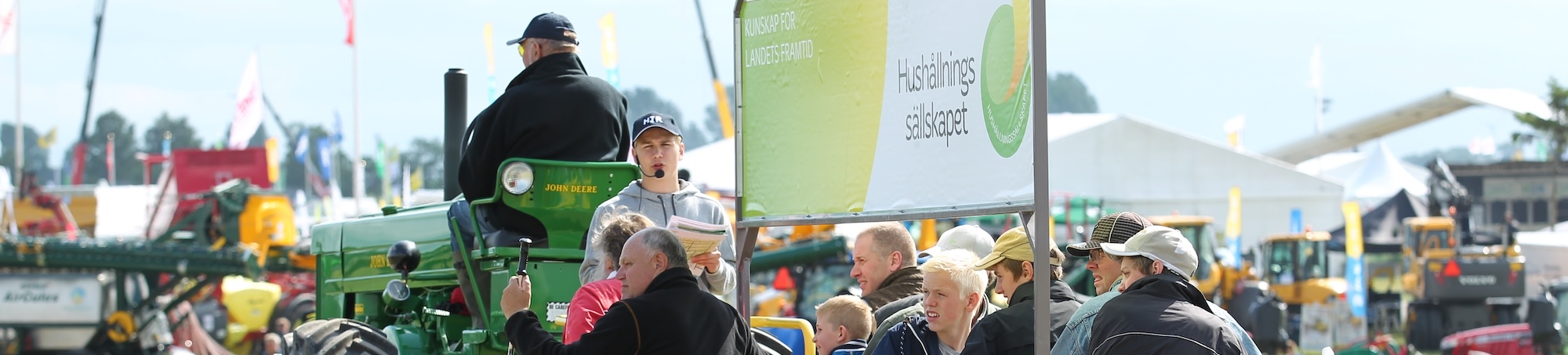 Man med mikrofon och papper står vid en grön John Deere-traktor och talar till en publik på en lantbruksmässa. Skylt i bakgrunden: ”KUNSKAP FÖR LANDETS FRAMTID” och ”Hushållnings sällskapet”.