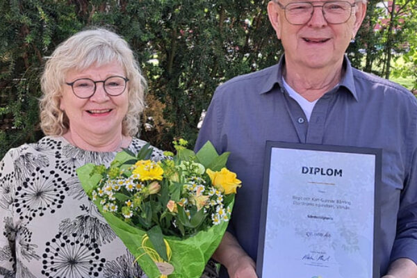Äldre par står utomhus framför grönska; mannen håller upp ett diplom och kvinnan ler med en bukett gula blommor. Diplomet har rubriken ”DIPLOM” med mindre text under, som inte är läsbar.