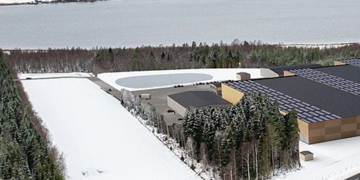 Aerial view shows a large industrial warehouse complex beside a snowy forest, with a plowed roadway in the foreground. Nearby are fenced lots and a round water basin, with a wide lake and scattered buildings beyond.