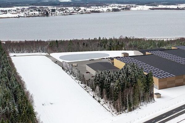 Aerial view shows a large industrial warehouse complex beside a snowy forest, with a plowed roadway in the foreground. Nearby are fenced lots and a round water basin, with a wide lake and scattered buildings beyond.