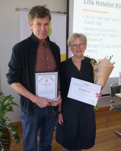 Two people pose indoors holding award documents; the woman also holds a bouquet. The certificate reads “VÄRDEBEVIS 10 000 kr,” with “Hushållningssällskapet Sjuhärad” visible, in front of a projected presentation slide.