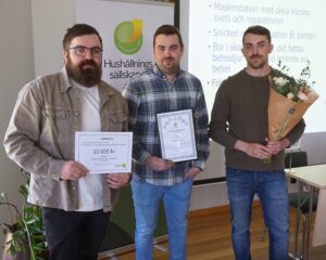 Three men pose indoors at an award presentation, holding a certificate, a framed diploma, and a bouquet. Behind them a banner reads “Hushållningssällskapet,” and the certificate shows “VÄRDEBEVIS 10 000 kr.”