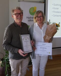 Two smiling people pose at an award event, holding a framed certificate and a bouquet with a “VÄRDEBEVIS” voucher reading “10 000 kr.” Behind them, a “Hushållningssällskapet” banner and a projected slide are visible.