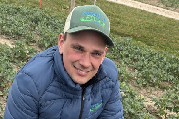 Leende man i blå vadderad jacka och grön keps med texten ”Borgaby Fältdagar 2023-09” hukar i en odling och tittar mot kameran; runt honom syns rader av gröna plantor och ett orange nätstaket.
