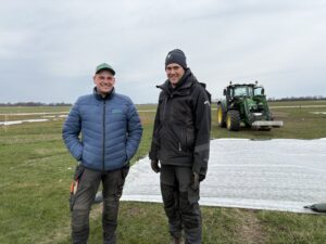 Två leende män i arbetskläder står på en åker och poserar framför en vit täckduk. I bakgrunden syns en grön traktor med lastare. Text på kläder: ”Borgeby Fältdagar”, ”Skåne”.