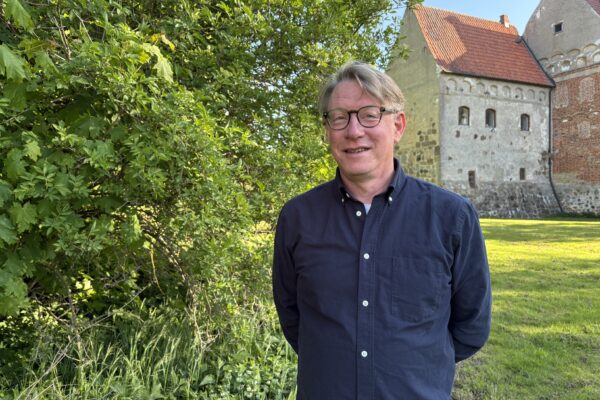 Leende man med glasögon står med händerna bakom ryggen vid en tät grön buske. I bakgrunden syns en äldre byggnad med grå murar och rött tegeltak i soligt väder.