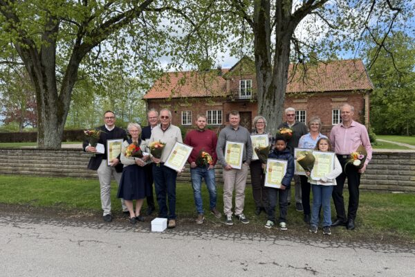 Grupp på elva personer poserar utomhus och håller buketter och inramade diplom med texten ”DIPLOM”. De står under stora träd framför en röd tegelbyggnad med tegeltak, som vid en prisutdelning.