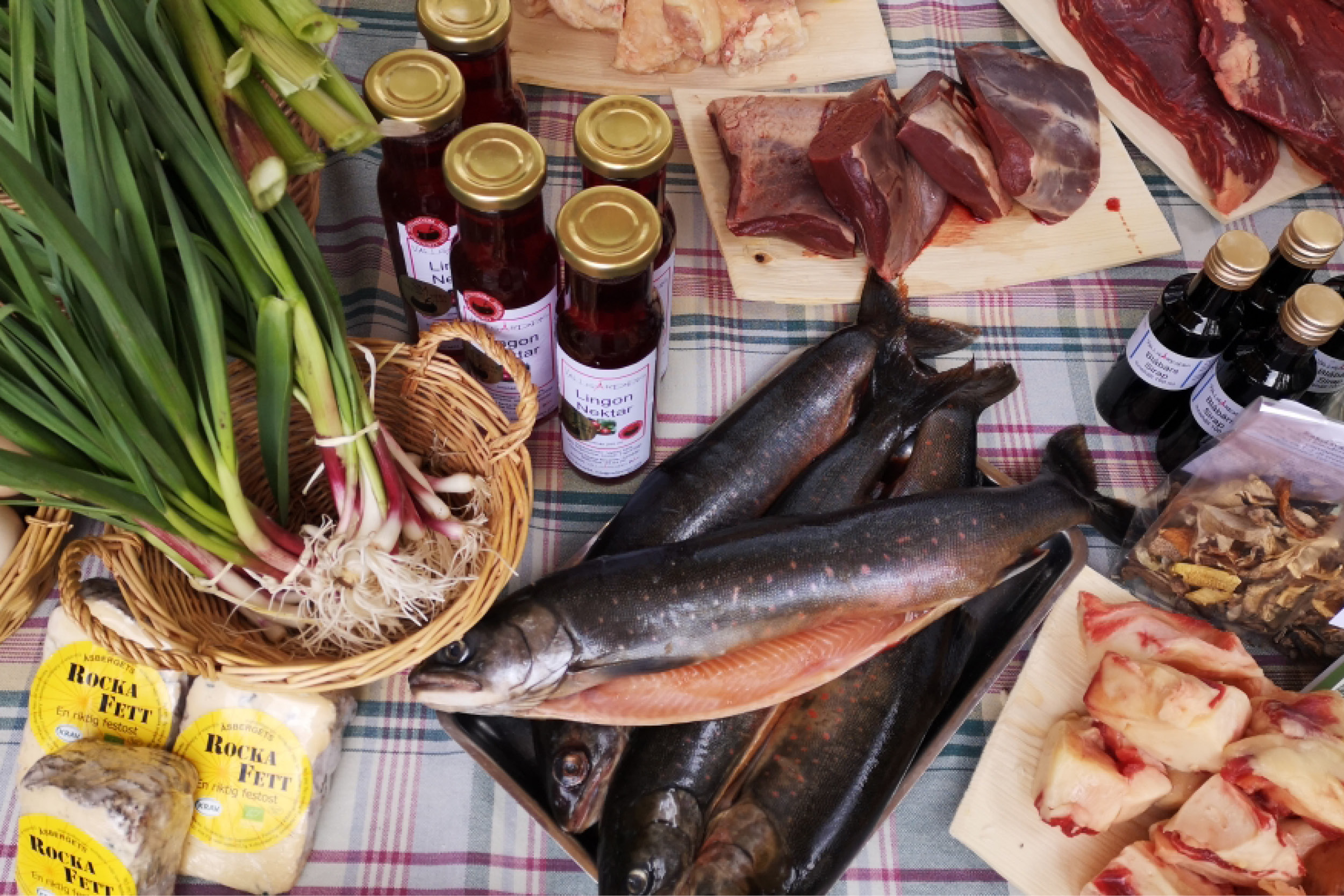Lokala matvaror ligger framdukade på en rutig duk: hela fiskar och fiskfilé på fat, styckat kött på skärbrädor, salladslök i korg, flaskor märkta ”Lingon NeKtar” samt förpackningar ”Rocka Fett”.