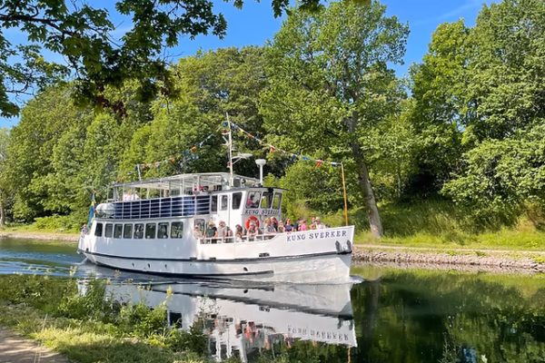 Vit passagerarbåt med texten ”KUNG SVERKER” glider fram på en stilla kanal, med resenärer på däck. Grönskande träd och blå himmel omger vattnet, där båten speglas nära strandkanten.