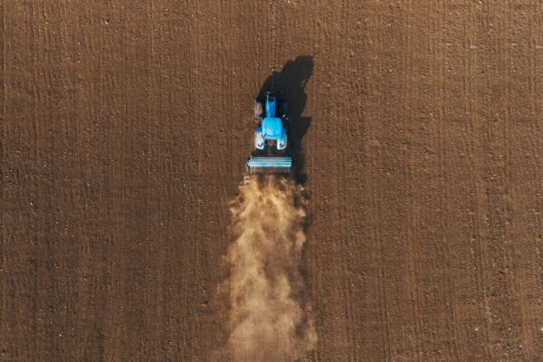 Blå traktor kör över en nyplöjd åker och drar en jordbearbetningsmaskin bakom sig, medan ett stort dammoln virvlar upp. Fotograferad rakt uppifrån, med tydliga raka spår i den bruna jorden.