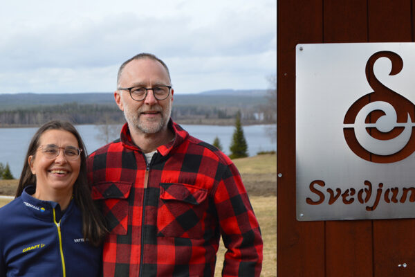 Två personer står leende utomhus med armen om varandra, bredvid en skylt på en byggnad. I bakgrunden syns sjö och skog. På skylten står “Svedjan Ost” med en stiliserad logotyp.