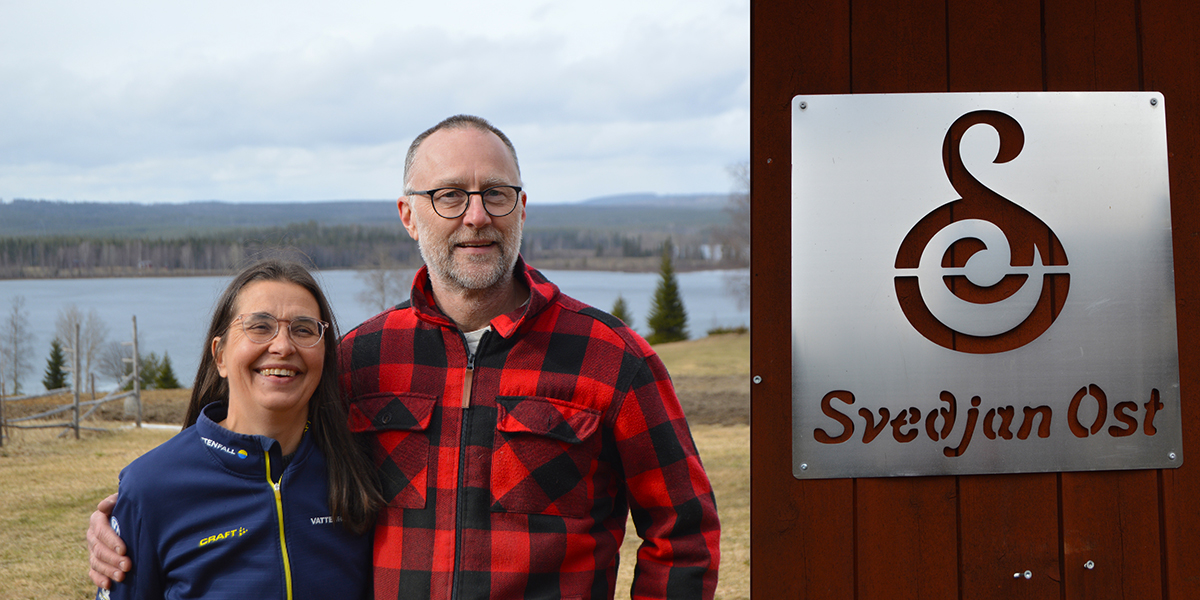 Två personer står leende utomhus med armen om varandra, bredvid en skylt på en byggnad. I bakgrunden syns sjö och skog. På skylten står “Svedjan Ost” med en stiliserad logotyp.
