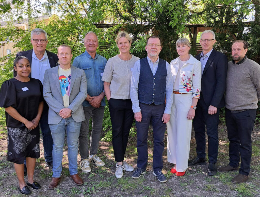Grupp på nio personer står uppställda och poserar utomhus bland träd och grönska, troligen en styrelsebild. De bär blandade kläder från kavaj till vardagsplagg och tittar mot kameran.